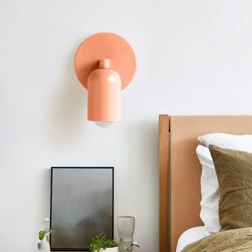 The Luna Glow modern wall light in pink, installed above a wooden bedside table in a contemporary bedroom setting. The light illuminates the space next to a bed with neutral bedding and decorative plants.