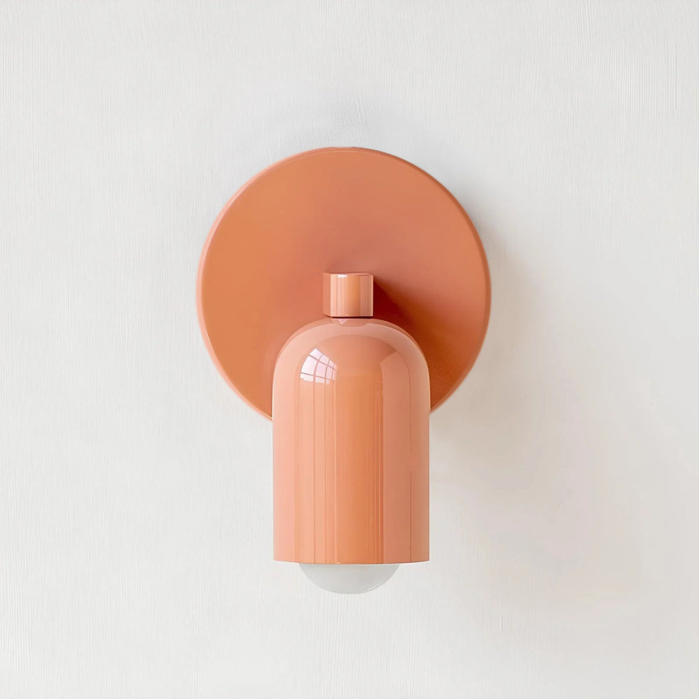 A close-up view of the Luna Glow modern wall light in a soft pink finish, featuring a cylindrical shade and a circular backplate, mounted on a clean white wall. Highlights its contemporary design and metal construction.