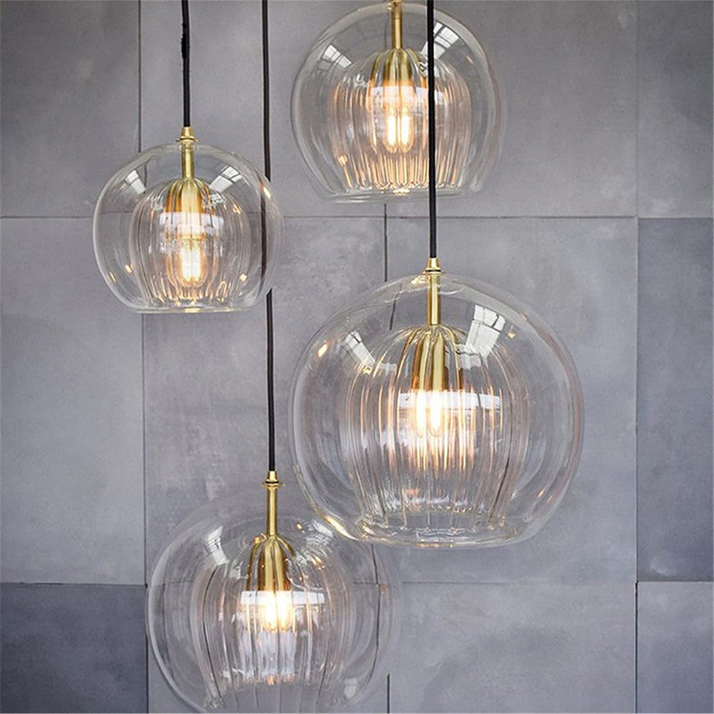Four Lustre Aura LED pendant lights in clear glass, hanging at different heights against a modern grey tiled wall, showcasing their design and warm glow.