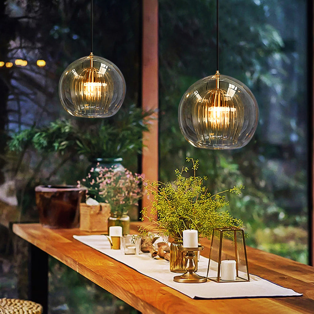 Two Lustre Aura LED pendant lights in clear glass illuminating a rustic wooden dining table decorated with plants and candles, set against a window with greenery.