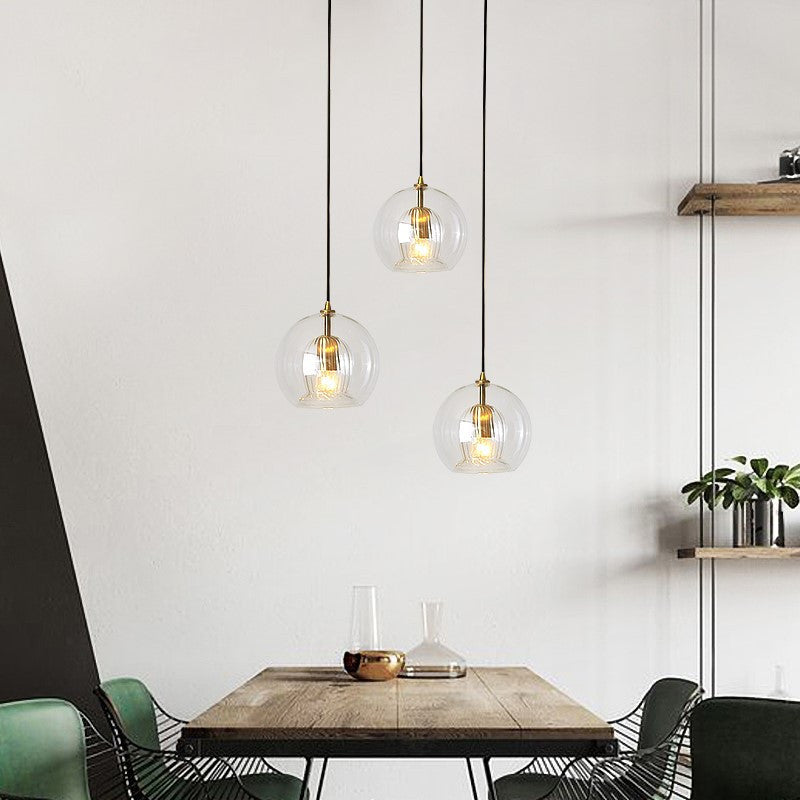 Three Lustre Aura LED pendant lights in clear glass with warm light, hanging at adjustable heights over a modern wooden dining table with green chairs, showcasing contemporary design and warm ambiance.