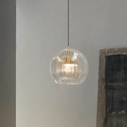 Lustre Aura LED pendant light featuring a clear outer glass globe and a ribbed inner clear glass shade with brass accents, emitting a warm glow. The modern adjustable height fixture hangs against a light gray wall, perfect for contemporary home decor.