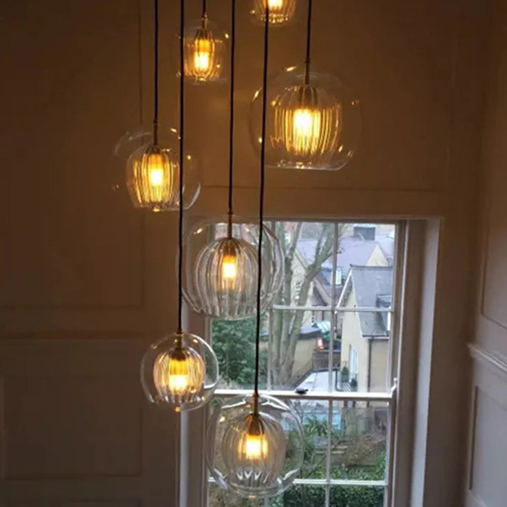 A collection of Lustre Aura LED pendant lights with clear amber glass and ribbed diffusers, emitting a warm glow, hanging at various adjustable heights in a stairwell next to a window.
