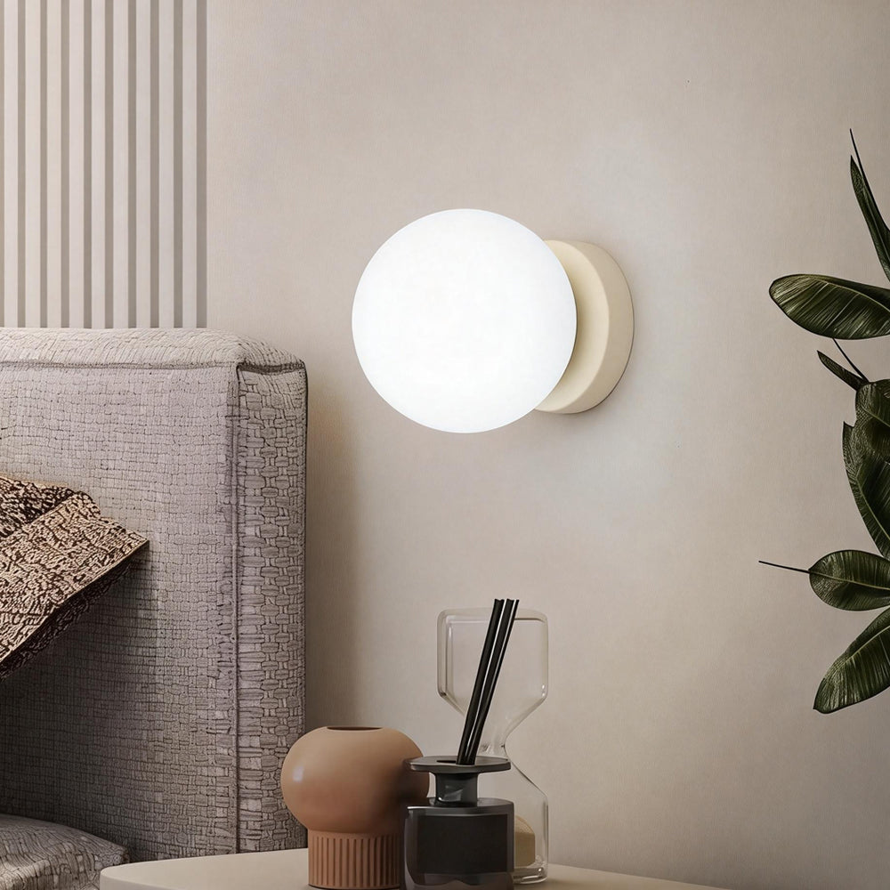 Luxe Aura beige iron glass wall lamp with LED illumination, glowing warmly on a light-colored wall in a modern bedroom setting, next to a bed with a textured headboard and a bedside table.
