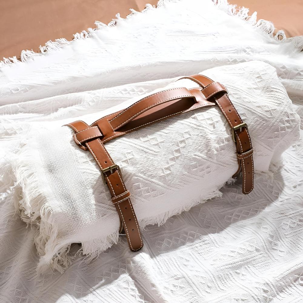 A rolled-up Cloud White Meadow Luxe picnic blanket with a water-resistant backing, secured by a stylish brown leather strap carrier, resting on a textured white surface. Highlights portability and design.