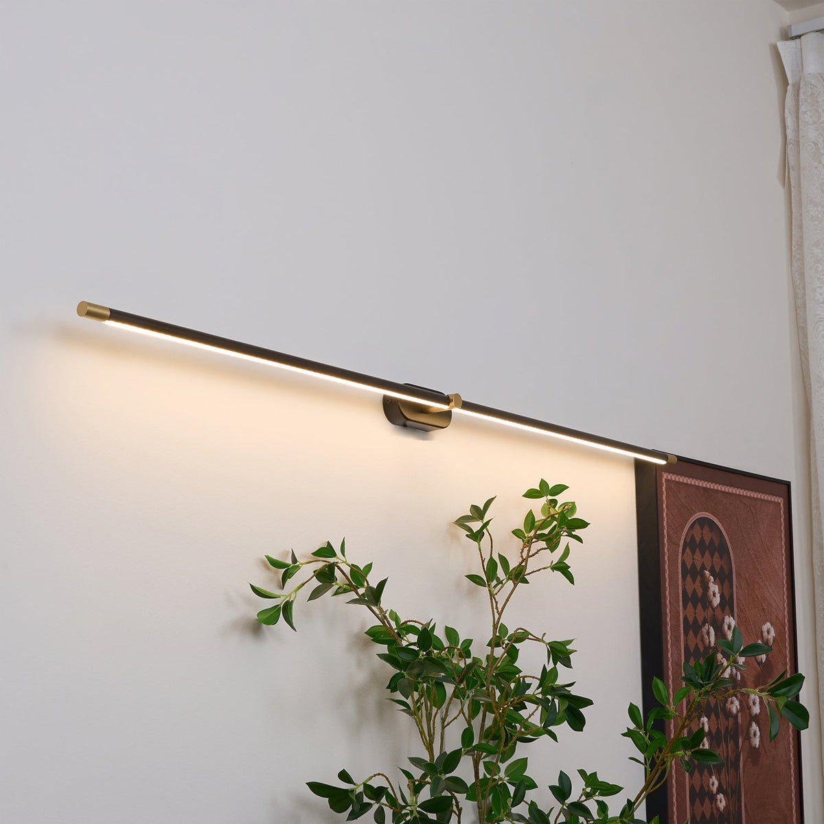 A black Vestra Minimalist Linear Rechargeable Wall Lamp with LED finish mounted on a white wall, casting warm light downwards. Below the lamp, a vibrant green plant adds a touch of nature, next to a framed artwork with an arched design.