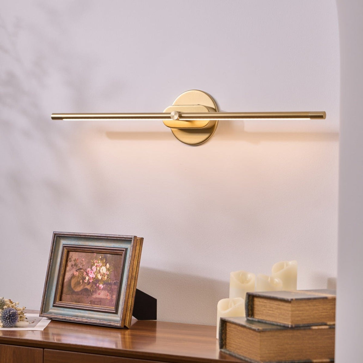 A gold Vestra Minimalist Linear Rechargeable Wall Lamp with LED finish mounted on a white wall, casting warm light downwards onto a wooden side table. The table holds a framed painting of flowers, a stack of vintage books, and several white pillar candles, creating a cozy ambiance.