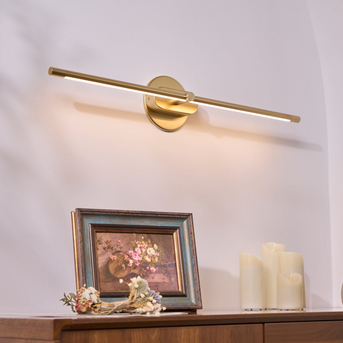 A gold Vestra Minimalist Linear Rechargeable Wall Lamp with LED finish mounted on a white wall, casting warm light downwards onto a wooden side table. The table features a framed painting of flowers and a small arrangement of dried flowers, enhancing the room's decor.