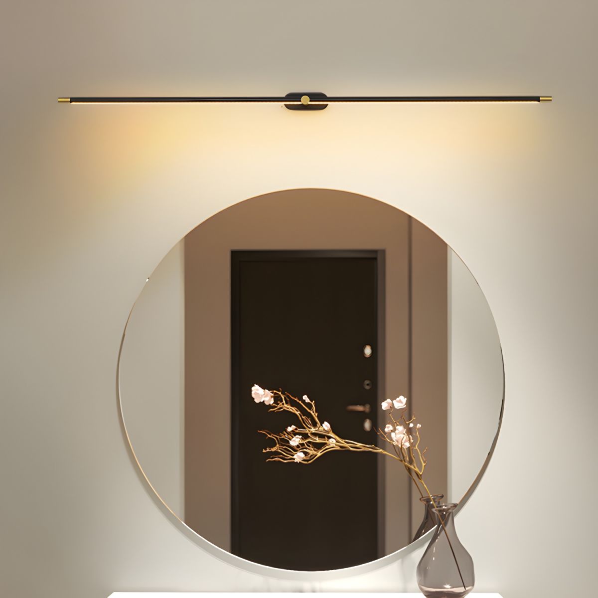 Vestra Minimalist Linear Rechargeable Wall Lamp in black, mounted above a round mirror with a vase of white flowers reflecting a doorway. The lamp emits a warm LED light.