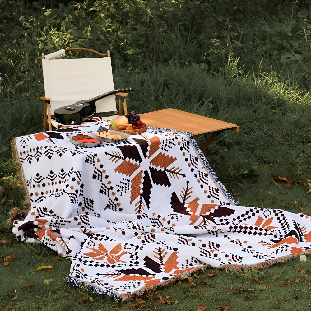 Nomad Oasis picnic blanket with Moroccan boho pattern partially draped over a small wooden table and spread on green grass, with a folding chair and ukulele in the background, highlighting its versatility.