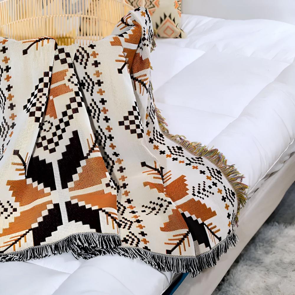 Nomad Oasis picnic blanket with Moroccan boho pattern folded and placed on a white bed with a rattan headboard, demonstrating its use as a stylish indoor throw blanket.