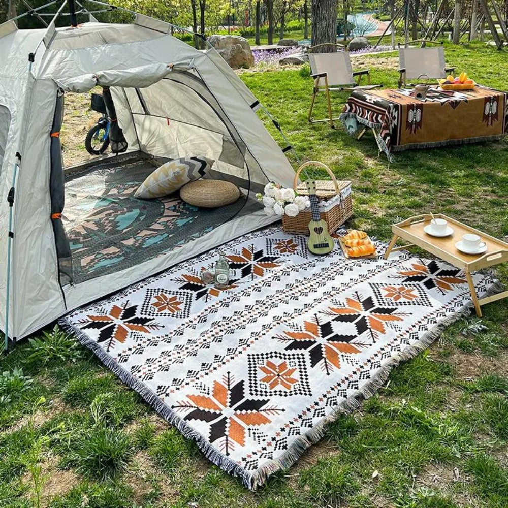 Nomad Oasis picnic blanket with Moroccan boho pattern laid out on green grass in front of a camping tent, surrounded by a picnic basket and food, perfect for outdoor adventures.