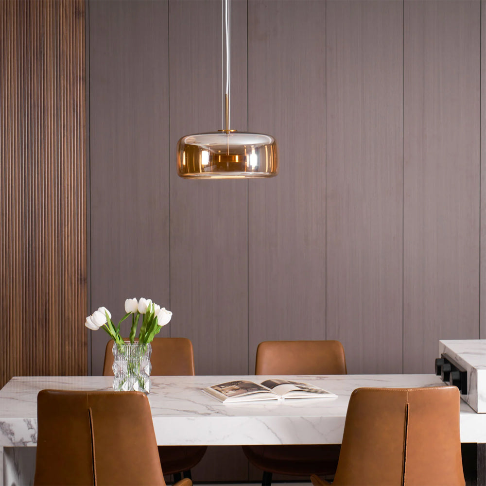 Nordic Glow Smoked Glass LED Pendant Light in amber, hanging above a modern marble dining table with brown leather chairs in a minimalist dining room. Features a contemporary design and warm ambient light.