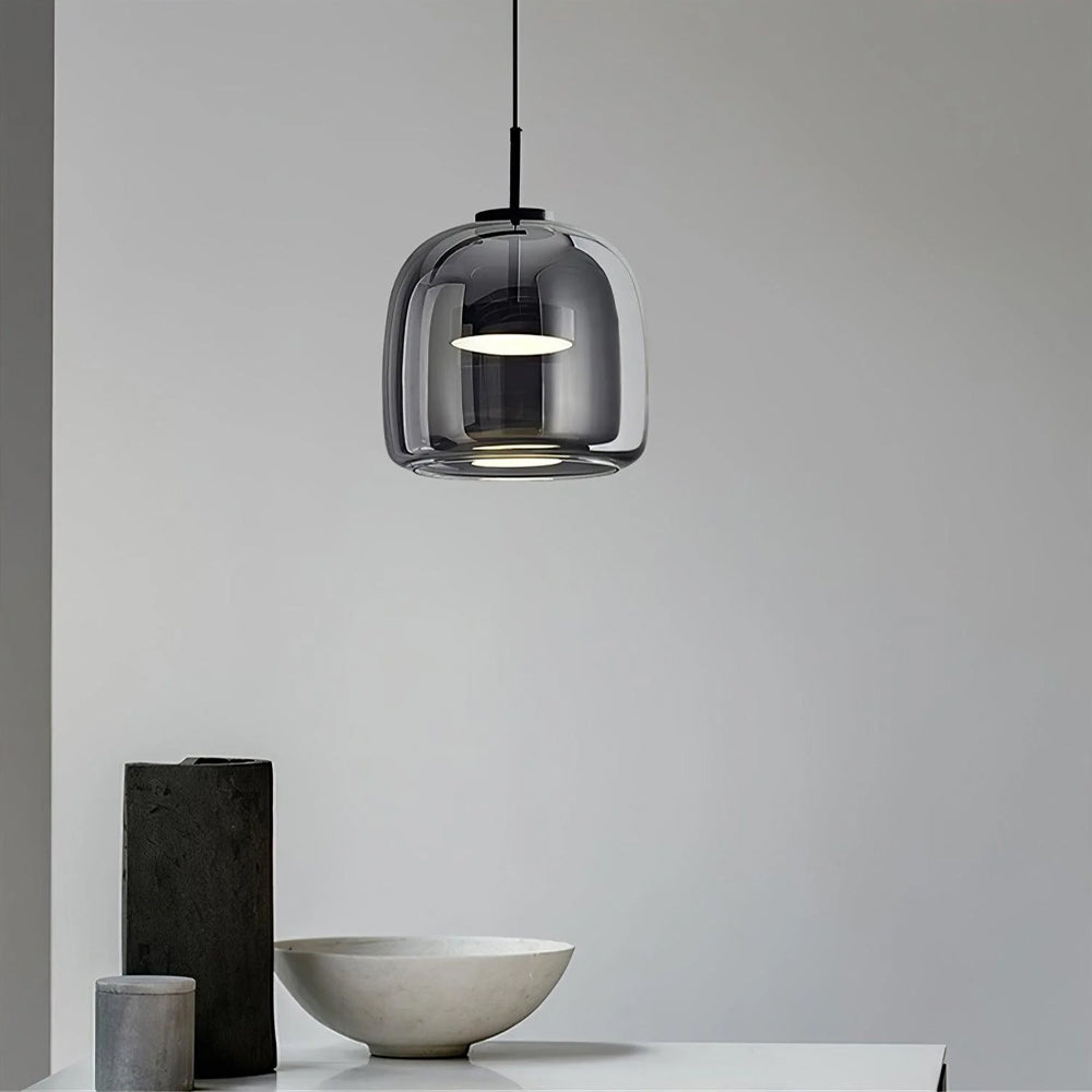 A single Nordic Glow smoked glass LED pendant light in grey, illuminated with a white light, hanging above a minimalist console table with a decorative bowl and vases, showcasing its contemporary design in a living space.
