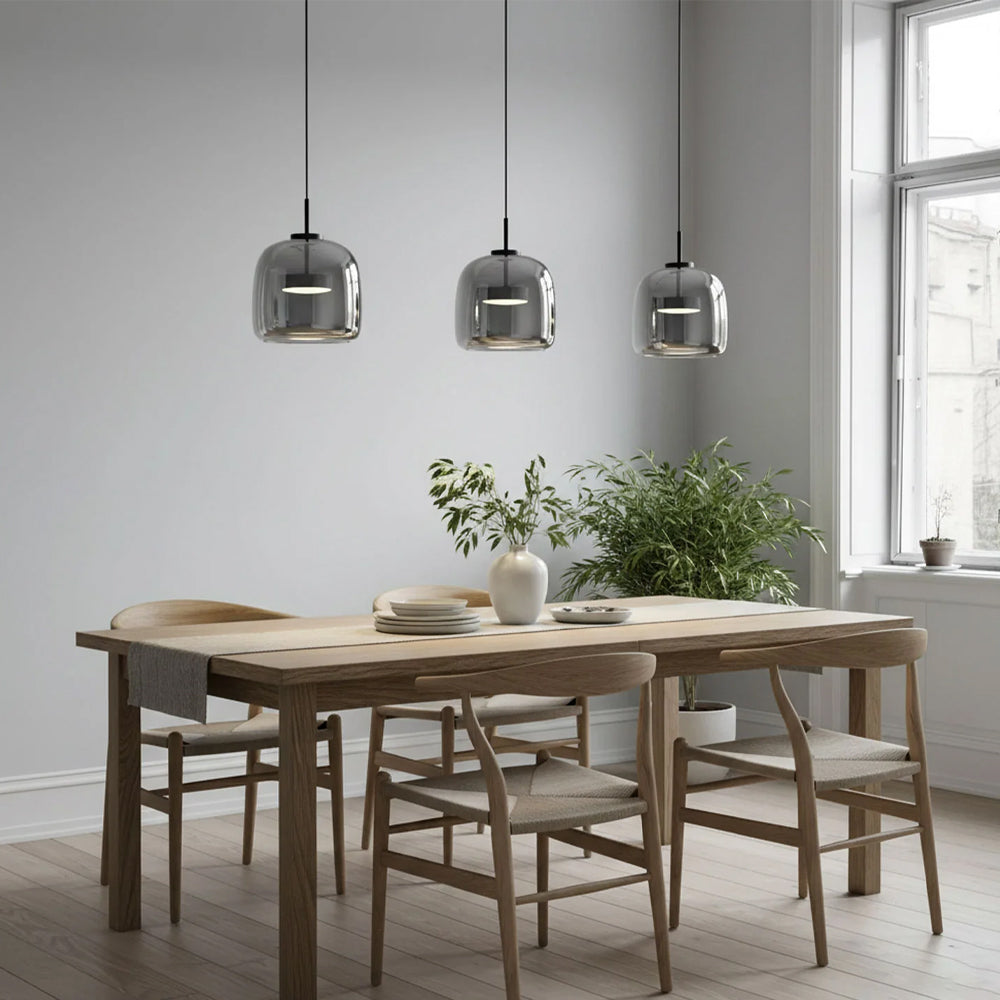 Three Nordic Glow smoked glass LED pendant lights in grey, hanging in a row above a wooden dining table with chairs, illuminating a modern dining room with natural light from a large window.