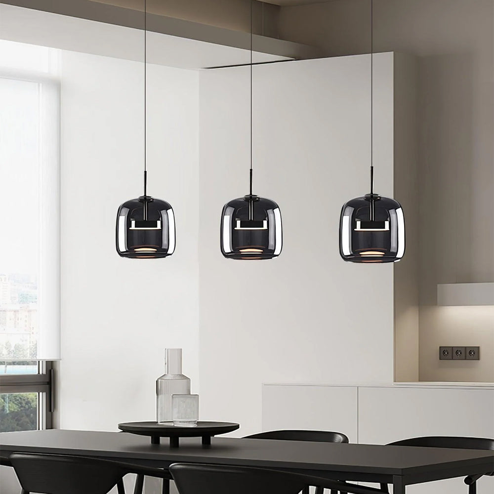 Three Nordic Glow smoked glass LED pendant lights in a darker grey finish, illuminated and hanging above a dark dining table in a contemporary kitchen or dining area. The industrial-style pendants add a modern touch to the space.