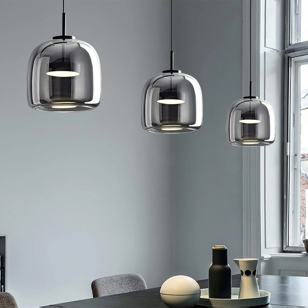 Three Nordic Glow smoked glass LED pendant lights in grey, illuminated with warm light, hanging above a modern dining table with minimalist decor. The lights feature a sleek, contemporary design, perfect for kitchen or dining room ambient lighting.