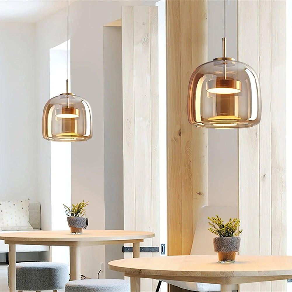 Two Nordic Glow smoked glass LED pendant lights in amber, emitting a warm glow, suspended above round wooden tables in a bright, minimalist cafe or dining setting. These modern hanging lights enhance the contemporary design.