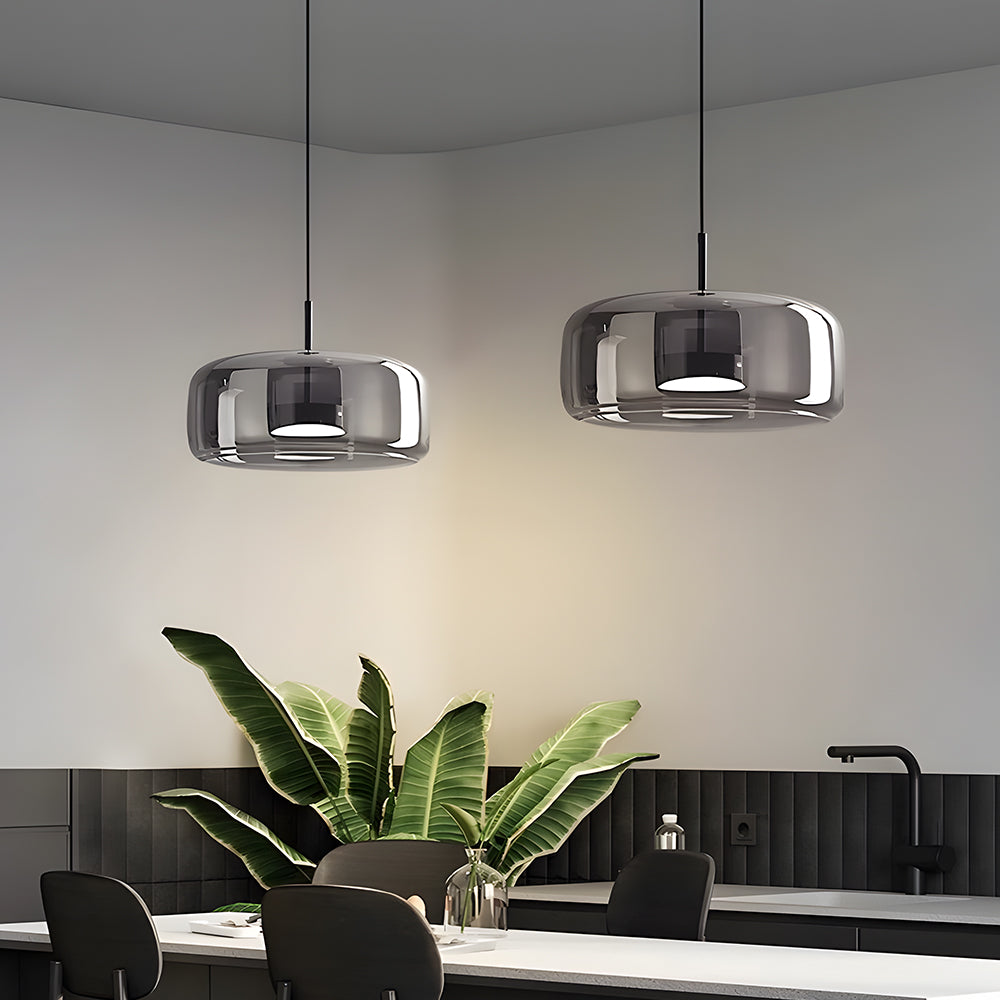 Two Nordic Glow B-shape grey smoked glass LED pendant lights with white light, hanging above a modern kitchen island or dining table with a large green plant, illustrating contemporary kitchen lighting.