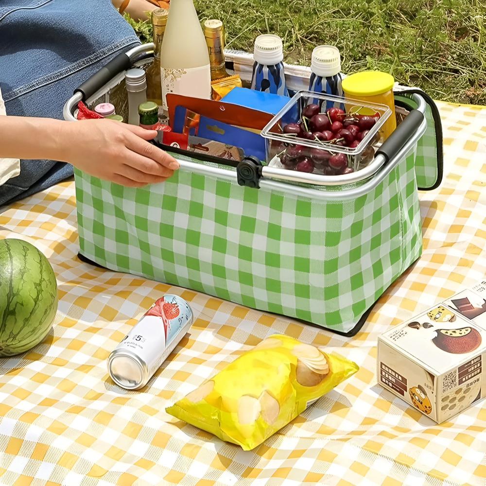 Green plaid checkered picnic basket, open and filled with cherries and bottles, set on a yellow checkered blanket on grass with a watermelon and snacks.