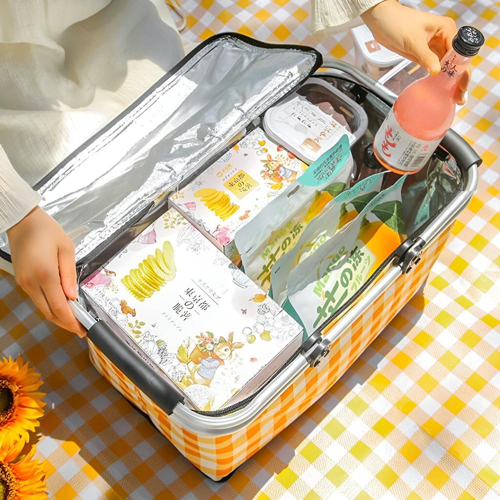 Yellow plaid checkered picnic basket, open and filled with various snacks and a pink drink, on a yellow checkered tablecloth with sunflowers. Hands are placing items inside.