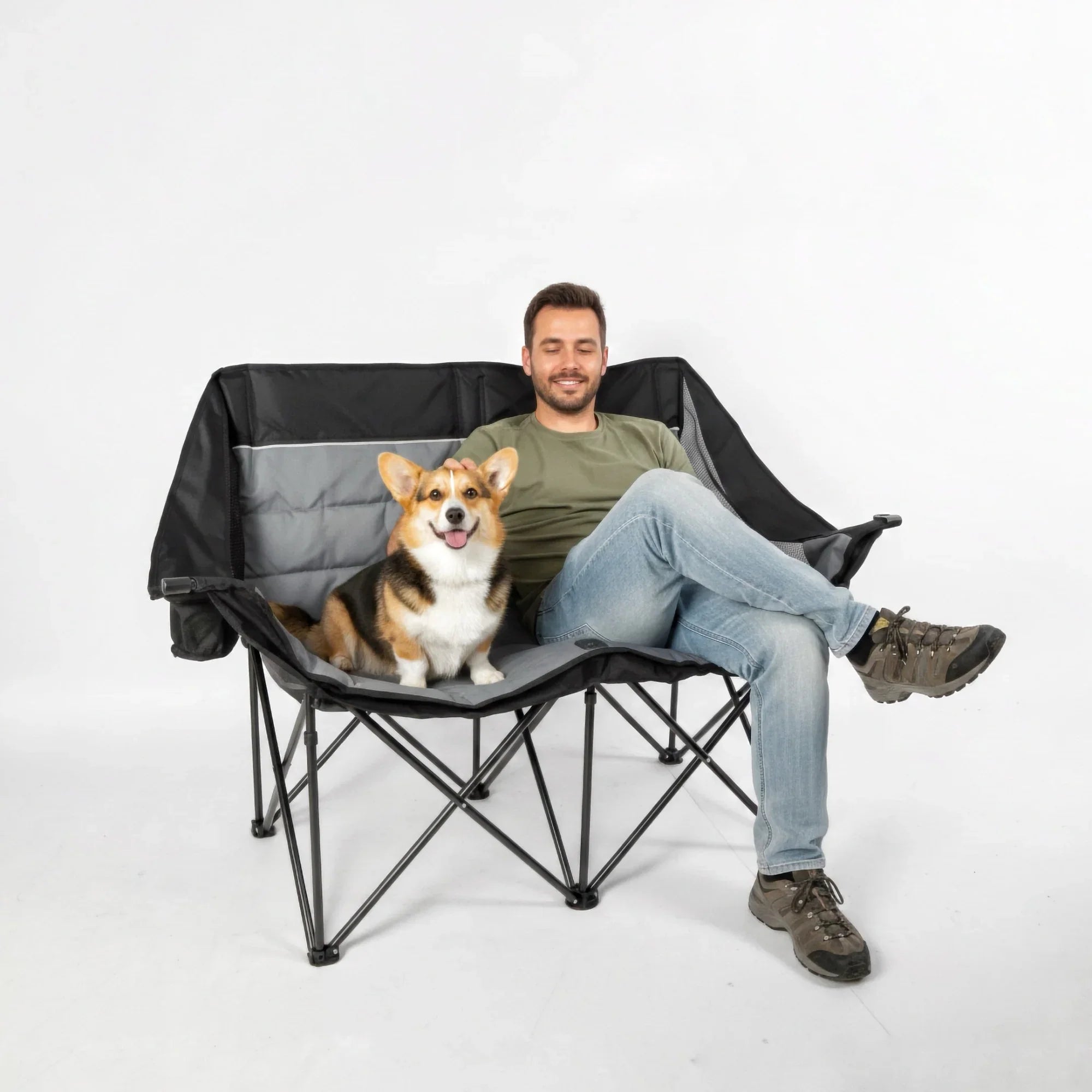 An oversized black and gray camping chair with hard arms, featuring a man and a corgi dog comfortably seated on it against a white background.