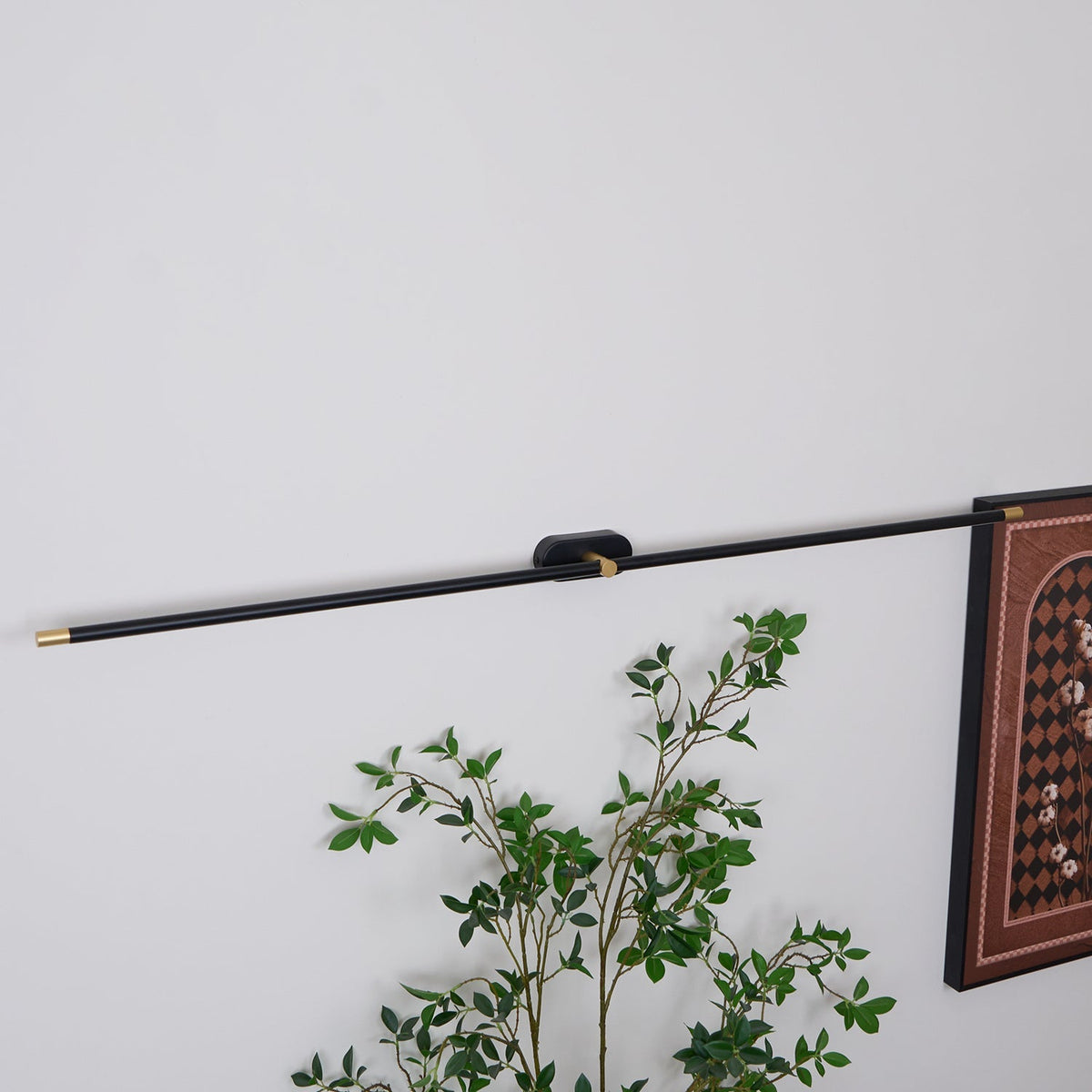 Vestra Minimalist Linear Rechargeable Wall Lamp in black metal with gold accents, installed on a white wall above a green plant, angled upwards, demonstrating its use in a modern living space.