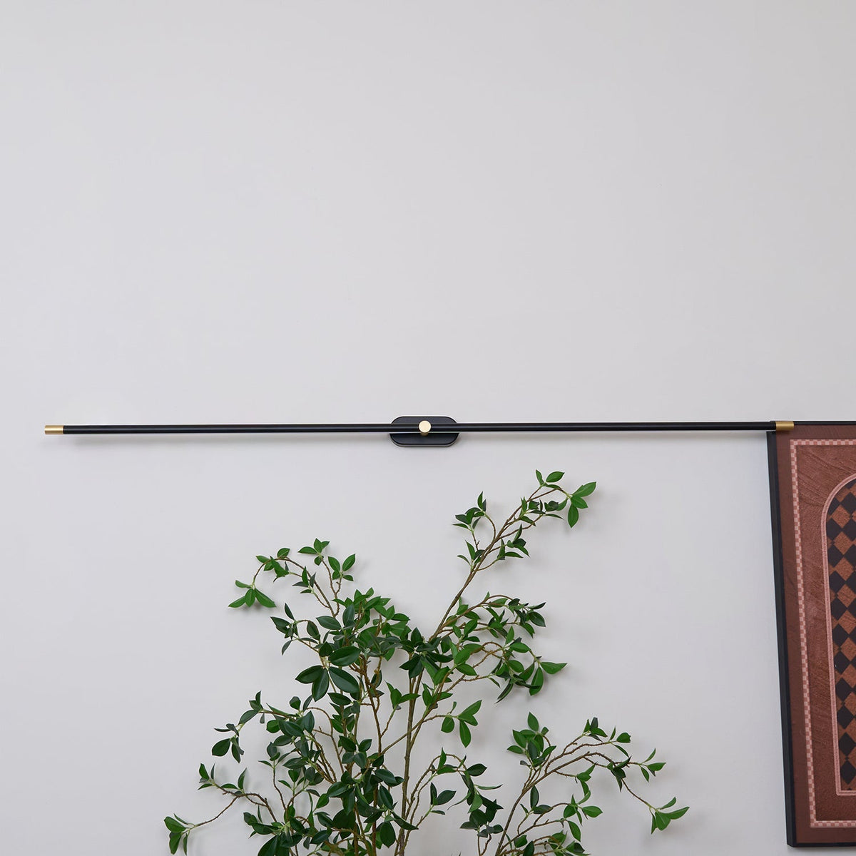 Vestra Minimalist Linear Rechargeable Wall Lamp in black metal with gold accents, installed horizontally on a white wall above a green plant, showcasing its sleek design in a contemporary setting.
