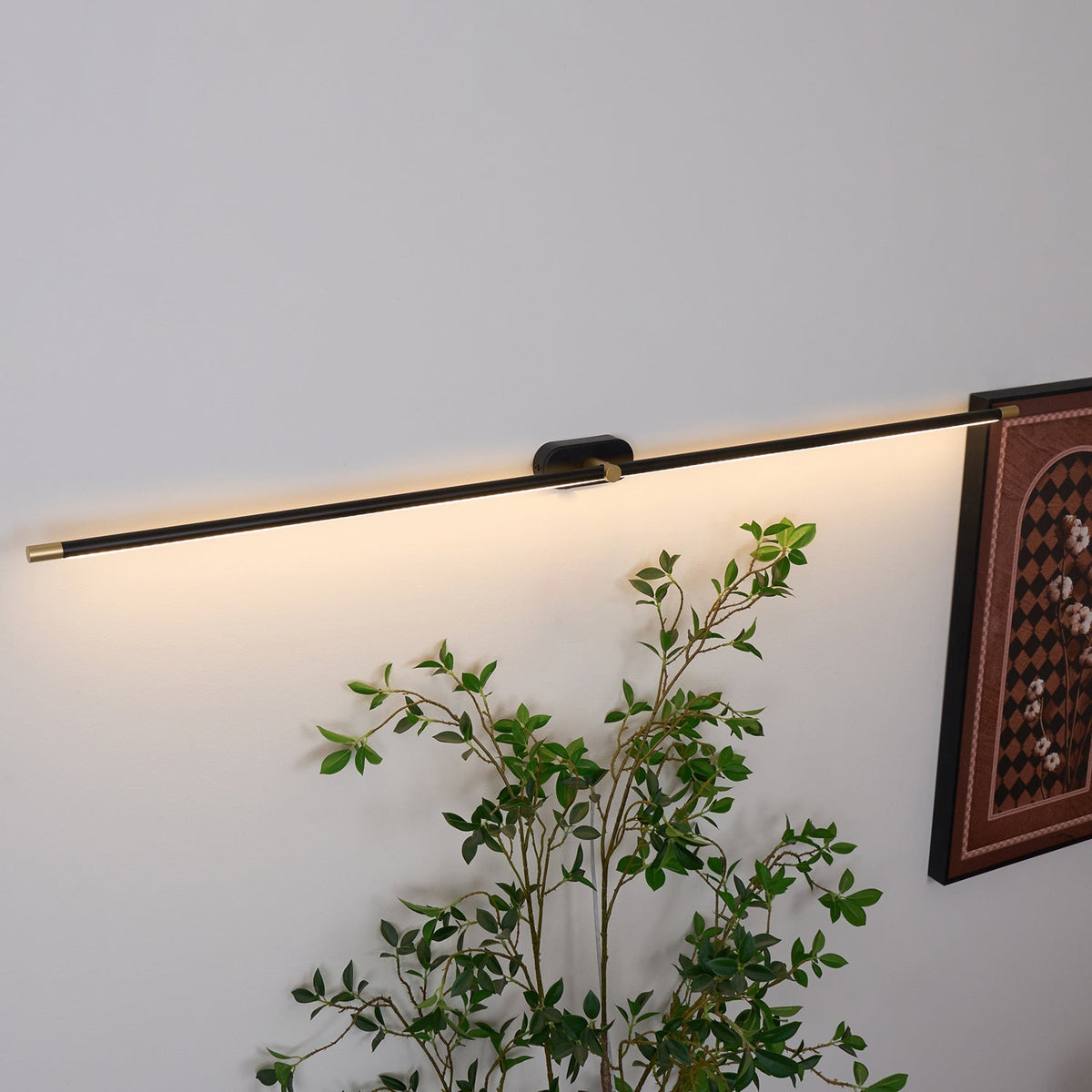 A Vestra Minimalist Linear Rechargeable Wall Lamp in black with gold accents, angled slightly upwards, emitting a warm LED light downwards, mounted on a white wall above a green plant. A framed artwork is partially visible.