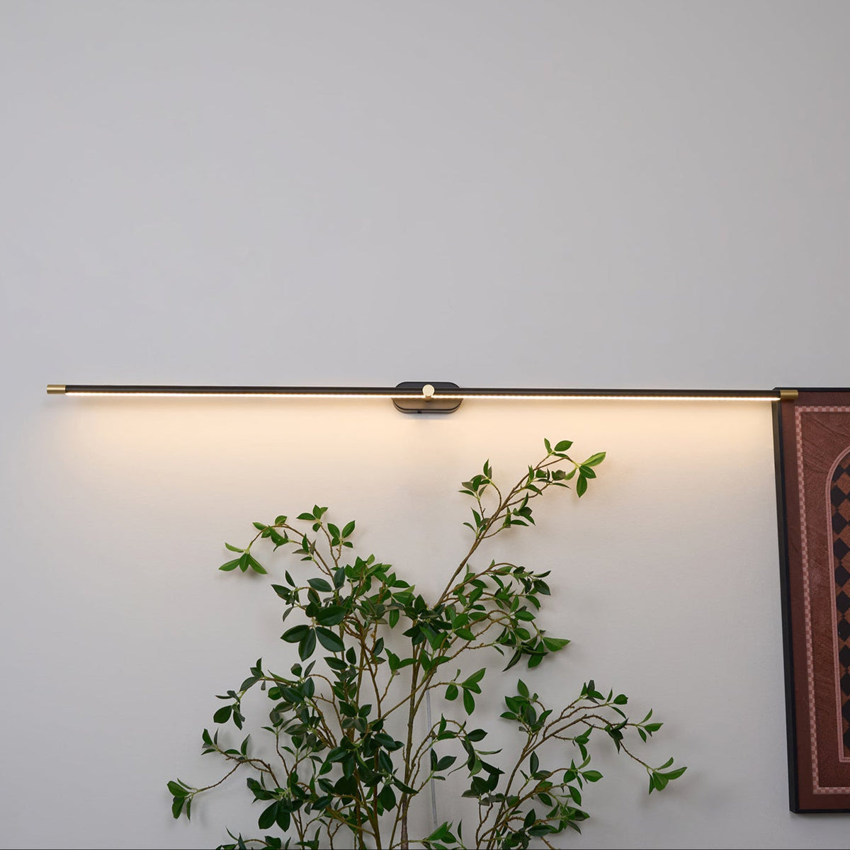 A Vestra Minimalist Linear Rechargeable Wall Lamp in black with gold accents, emitting a warm LED light downwards, mounted on a white wall above a green plant. A framed artwork is partially visible on the right.