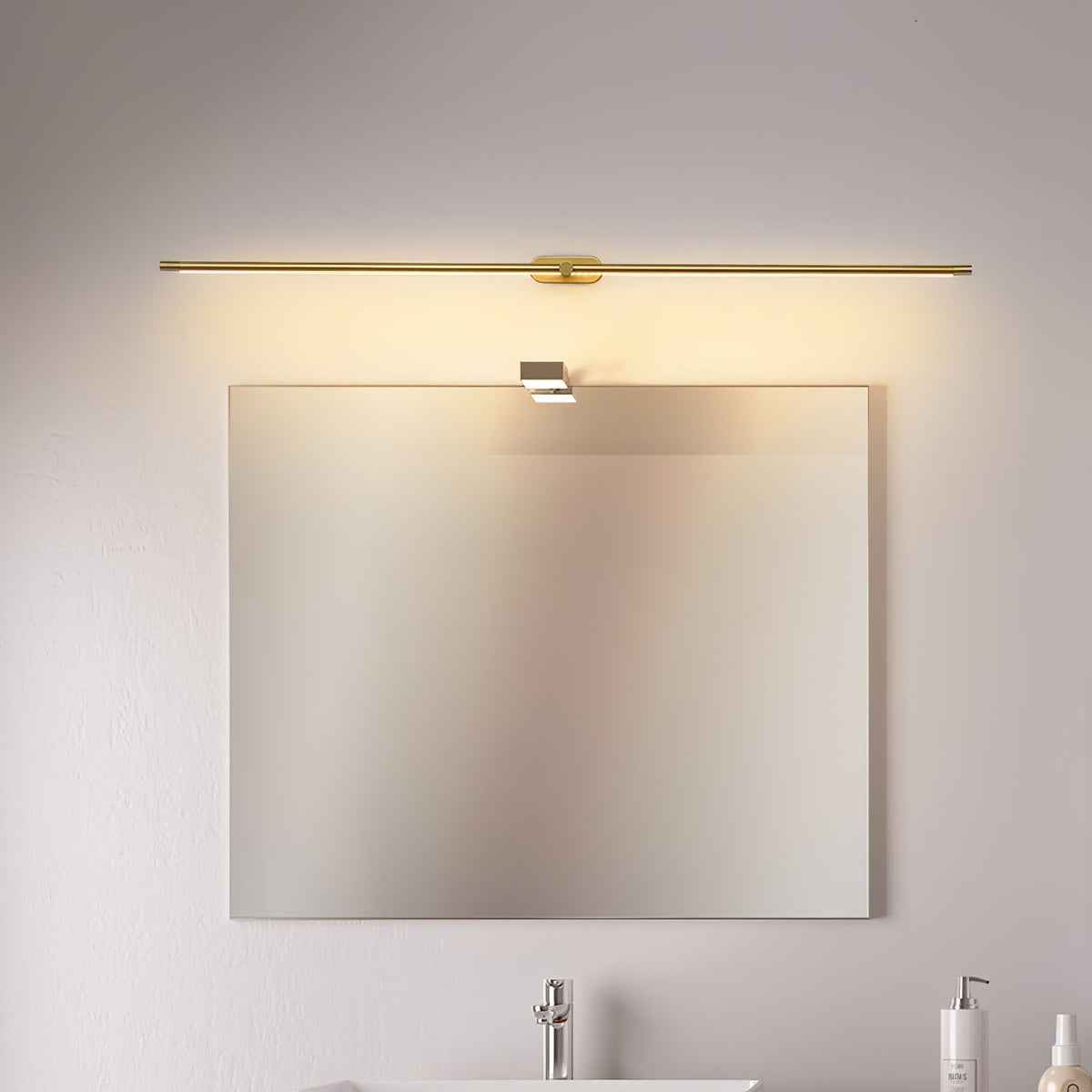 A Vestra Minimalist Linear Rechargeable Wall Lamp in gold, mounted above a rectangular mirror in a modern bathroom setting, illuminating a sink and faucet with warm LED light.