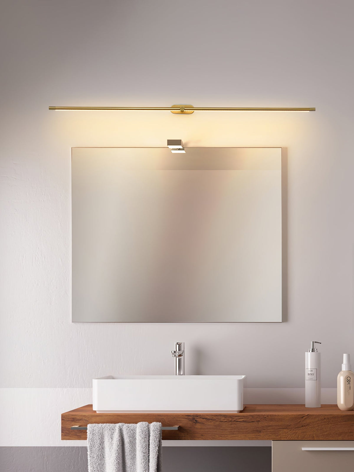 A Vestra Minimalist Linear Rechargeable Wall Lamp in gold, mounted horizontally above a square mirror in a bathroom, illuminating a white sink, wooden countertop, and folded towel.