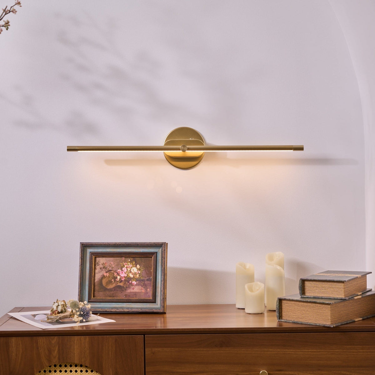 Vestra Minimalist Linear Rechargeable Wall Lamp Metal LED Finish in gold, illuminated, mounted above a wooden dresser with a framed floral picture and candles.
