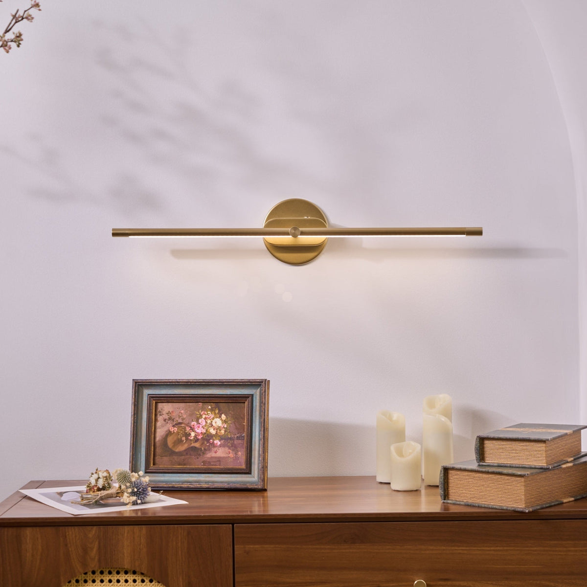 Vestra Minimalist Linear Rechargeable Wall Lamp Metal LED Finish in gold, illuminated, mounted above a wooden dresser with a framed floral picture and candles.